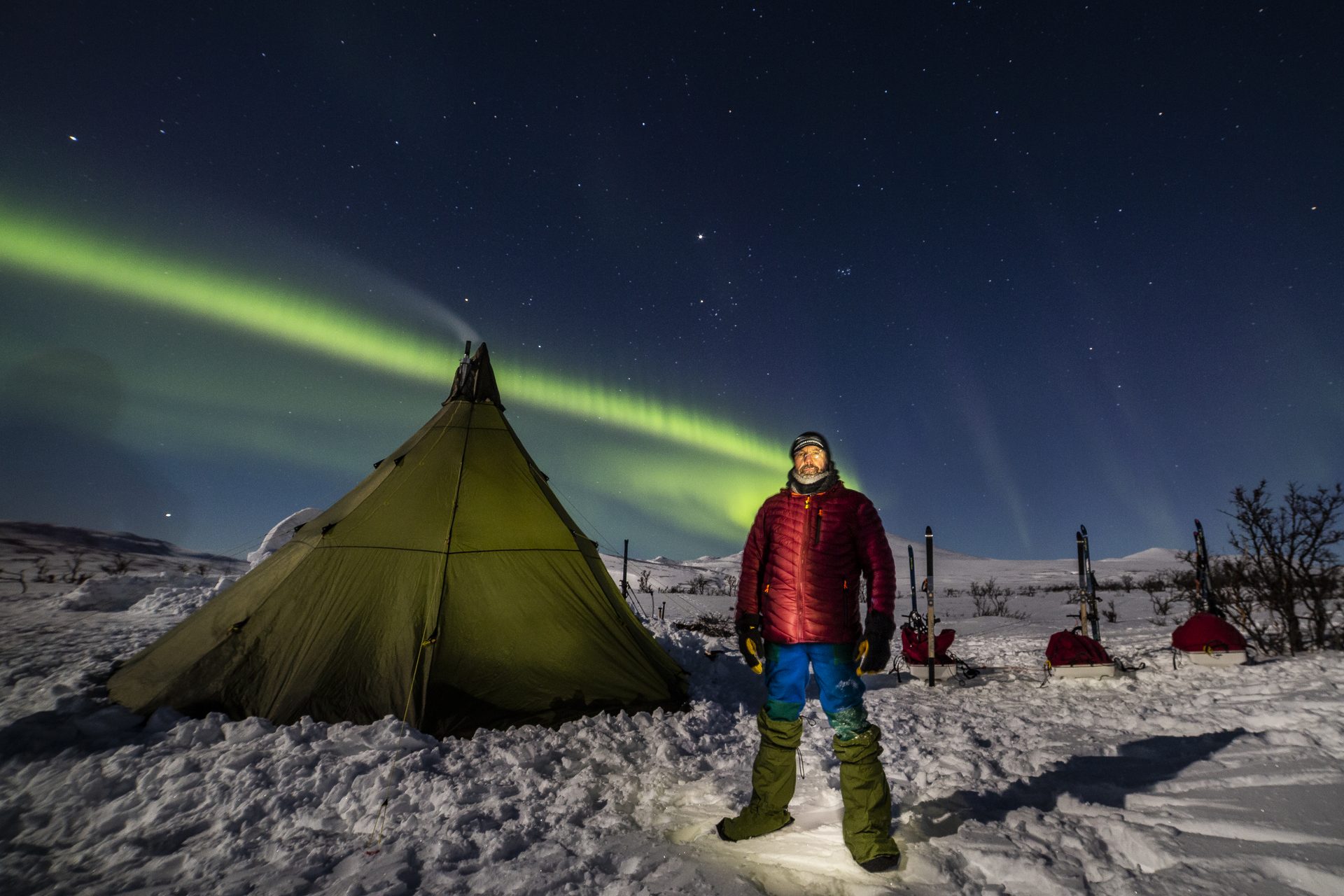 Expedicionario junto al tipi de campamento bajo una aurora boreal verde en el macizo del Kebnekaise